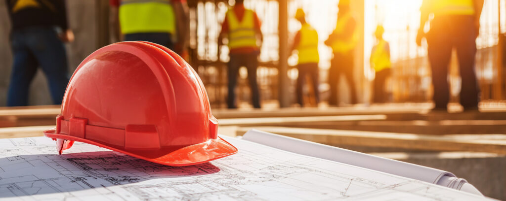 A red hard hat rests on construction blueprints, symbolizing safety and planning in bustling construction site. Workers in safety vests are visible in background, emphasizing teamwork and diligence.